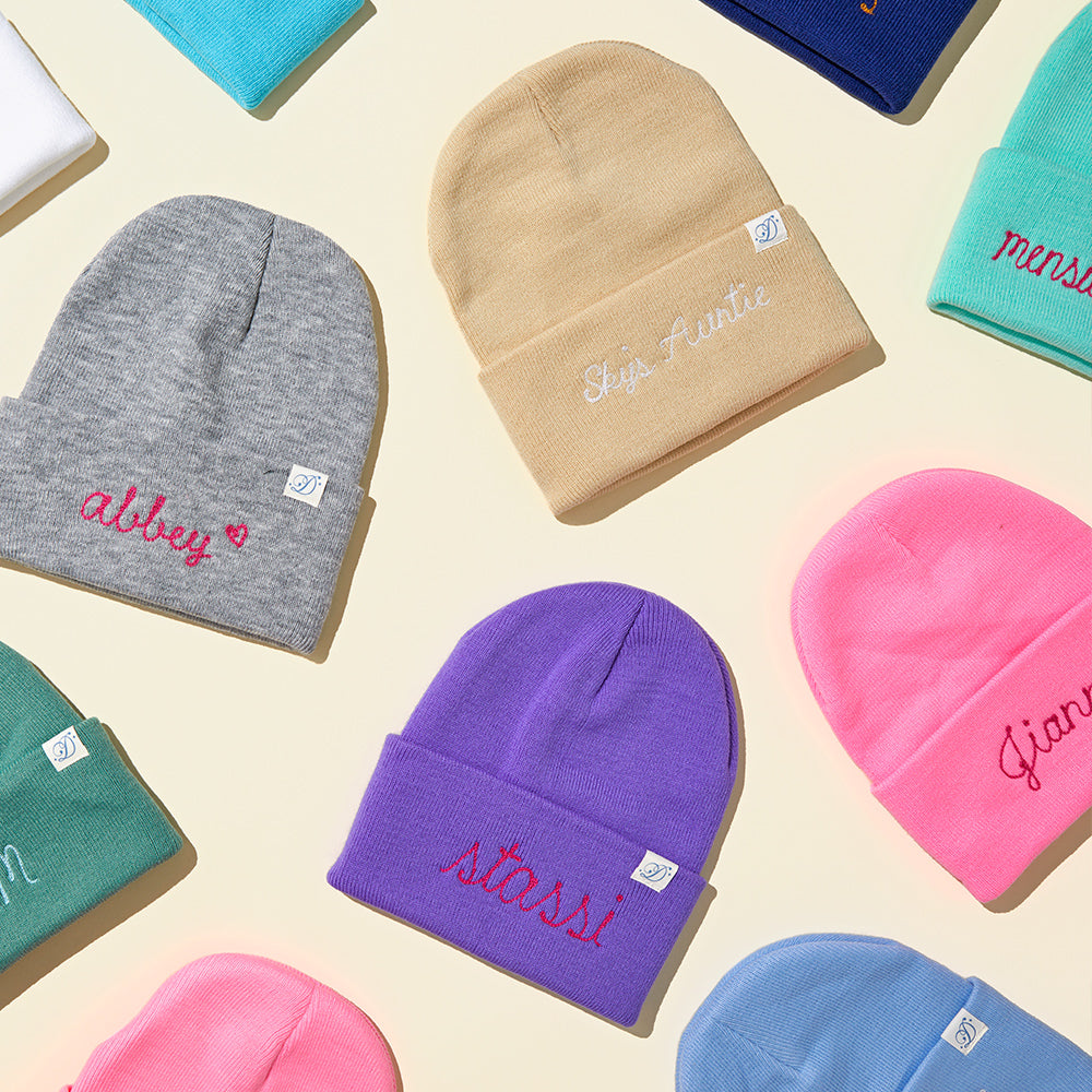 Variety of colorful beanies available for personalization with names, displayed on a wooden surface, showcasing the unique chainstitch embroidery. Daily Disco Beanies